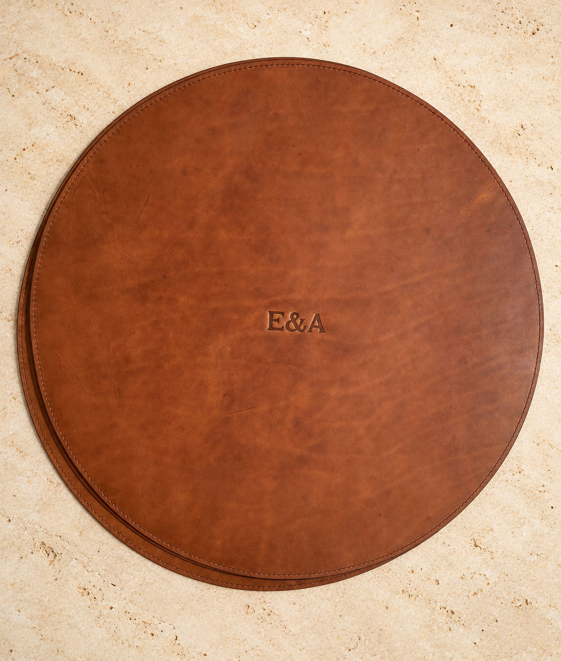 Brown circular leather mat personalized with debossed E&A initials on warm stone