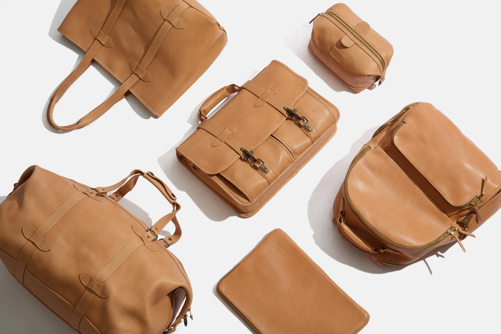 A coordinated set of tan leather travel and office bags arranged on a white background, including a tote, briefcase, backpack, duffel, and two small zip pouches. #color_essex-natural
