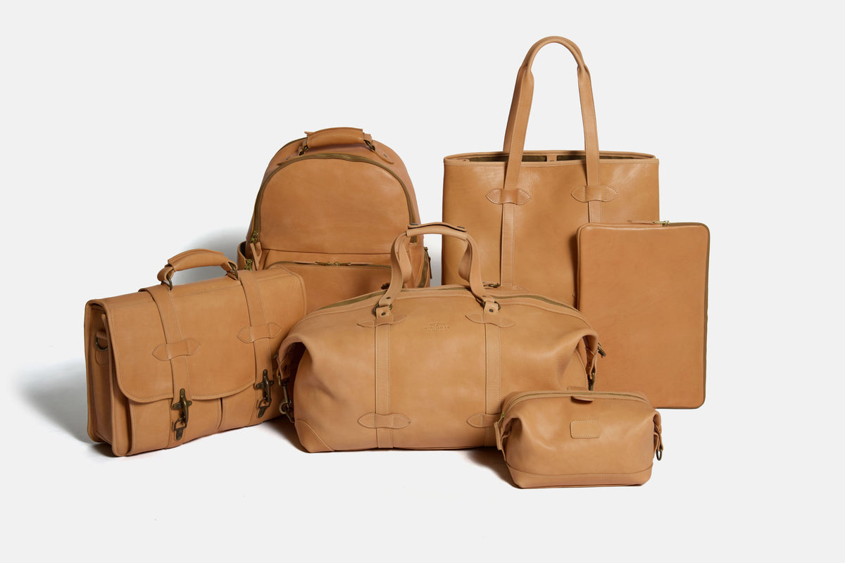 A collection of tan leather travel and luggage accessories arranged together on a light gray background, including a duffel bag, backpack, tote bag, briefcase, laptop sleeve, and small pouch. #color_essex-natural